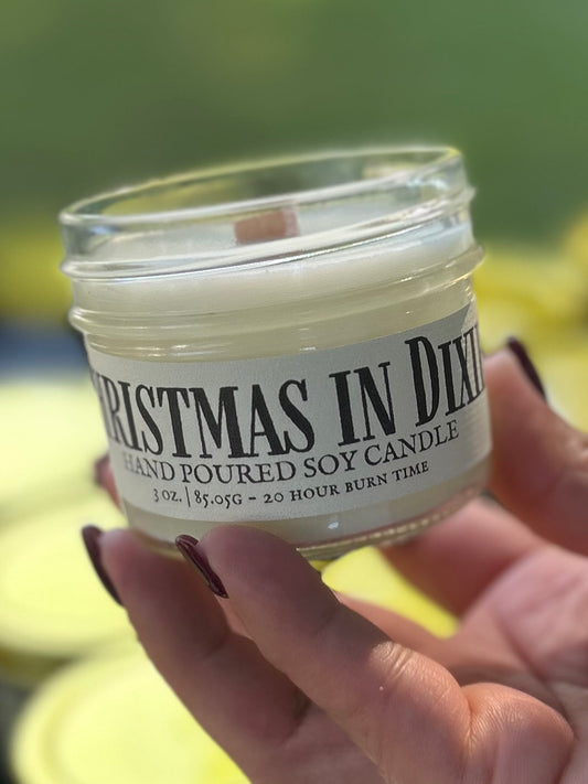 Hand holding a small candle jar labeled 'Christmas in Dixie' with a blurred background.